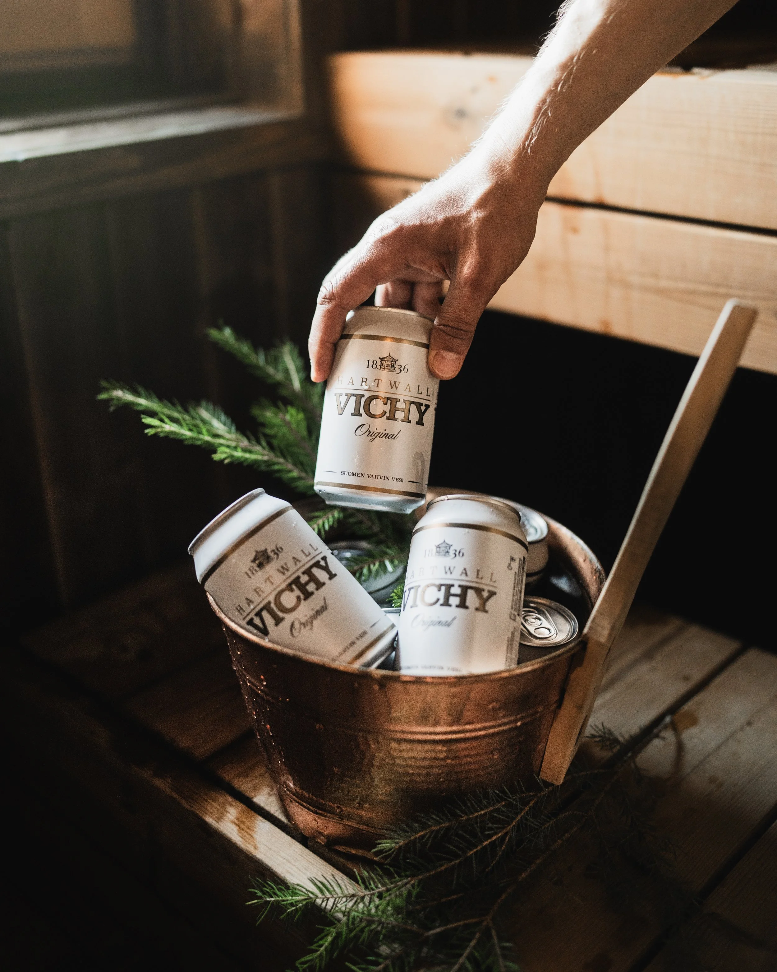 Sauna iA person's hand is reaching into a copper bucket with white cans of Vichy Original mineral water. Some cans are upright, and some are tilted inside the bucket, which is decorated with evergreen branches, on a wooden surface near a wooden wall.